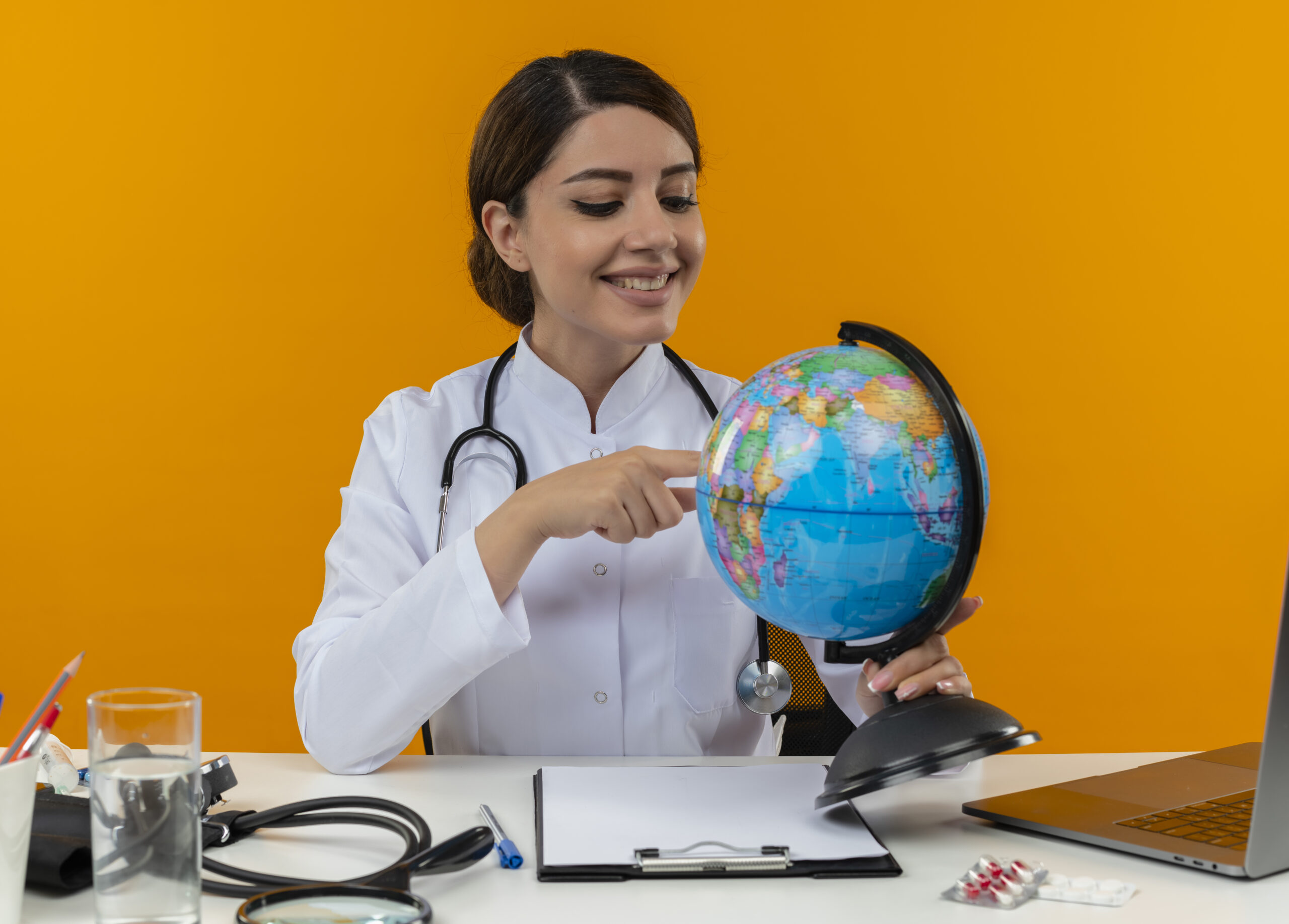 smiling young female doctor wearing medical robe with stethoscope sitting at desk work on computer with medical tools holding and putting finger on globe on isolation yellow background